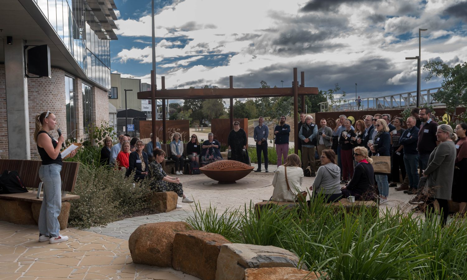 Image of a large group of people watching a speaker in a garden setting outdoors. Some are seated, some are standing. Sky is cloudy and moody.