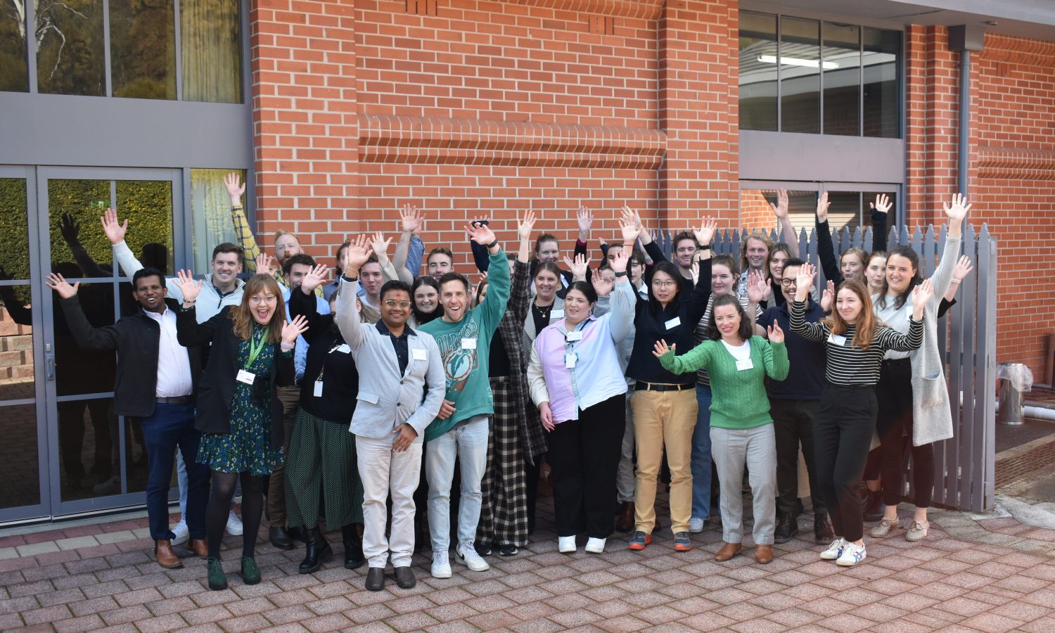 Large group photo outdoors in front of a red brick building. People smiling and waving in casual business attire.