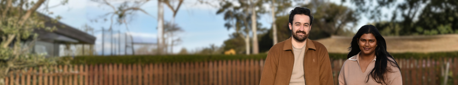 image shows a young man with a beard and a woman with long dark hair sitting facing the front. there is a lawn and picket fence leading into a bush landscape behind them